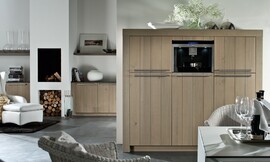 Hier ziet u een detailfoto van de hoogkast/apparatuurkast met brede wangen, stanggrepen en verticale strepen in het hout. Ook ziet u hoe hetzelfde concept is doorgevoerd in de woonkamer. Aparte eettafel met rieten stoelen. Zuordnung: Stil Landelijke keukens, Planungsart Detail keukenontwerp