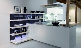 Deze open keuken heeft een gereduceerde look met witte fronten en een dun, metallic aanrechtblad. Het kookeiland staat haaks op een open wandgedeelte. Deze open wand met legplanken is ideaal om alles direct bij de hand te hebben, maar kan tegelijkertijd ook decoratief worden gebruikt. De blauwe achtergrond van de open kast zorgt voor een speels accent. Op deze open wandkast sluit het keukenblok met de spoelbak aan. De onder- en bovenkasten bieden voldoende bergruimte. Een keukenplanning zoals deze is ook erg geschikt voor grote ruimtes die deels als woonkamer functioneren. Zuordnung: Stil Design-keukens, Planungsart Open keuken (woonkeuken)