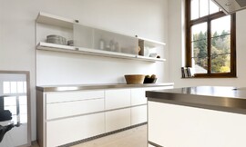 Deze moderne witte designkeuken maakt met zijn roestvrijstalen aanrechtbladen en greeploze witte fronten een strakke indruk. Aan de wand is een keukenblok geplaatst met grote, praktische lades. De lage kast daarboven heeft verschuifbare matglazen fronten en is voor een deel open. Het kookeiland heeft hetzelfde design: roestvrijstalen werkblad, witte greeploze fronten. Niets verstoort hier de kalmte die deze keuken uitstraalt. Zuordnung: Stil Luxe keukens, Planungsart keukenblok