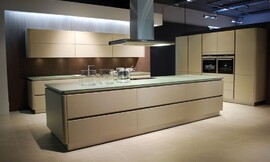 Ruime eilandkeuken met kookveld op het kookeiland. Beige fronten met greeplijsten, afgewerkt met rondingen. Dun glazen aanrechtblad, kleurtint richting groen. Zuordnung: Stil Design-keukens, Planungsart Keuken met keukeneiland